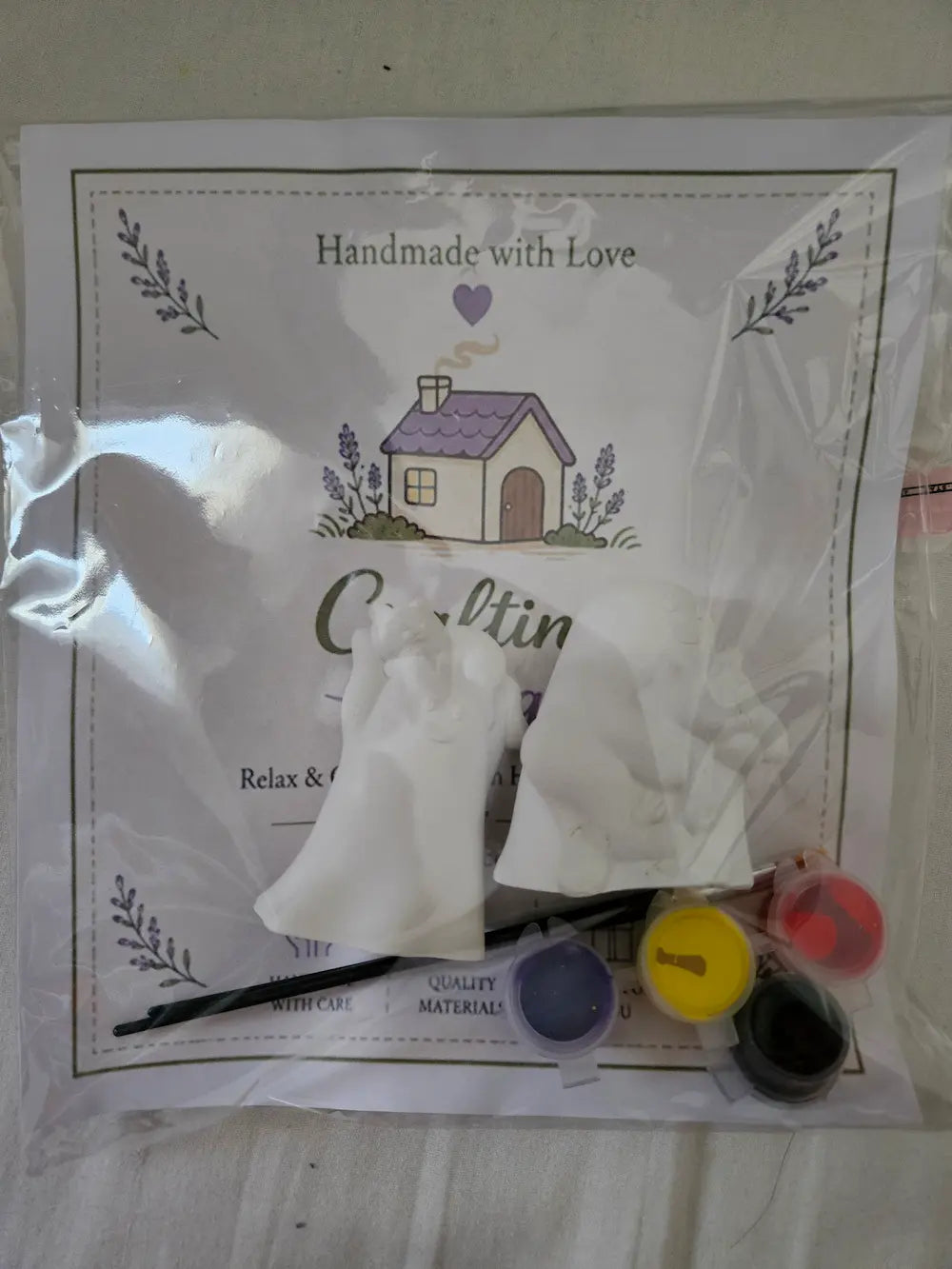 A packaged DIY craft kit containing white fabric gloves, a paintbrush, and several small paint pots in purple, yellow, red, and black, all resting on a printed white card that reads 'Handmade with Love' and 'Crafting'.