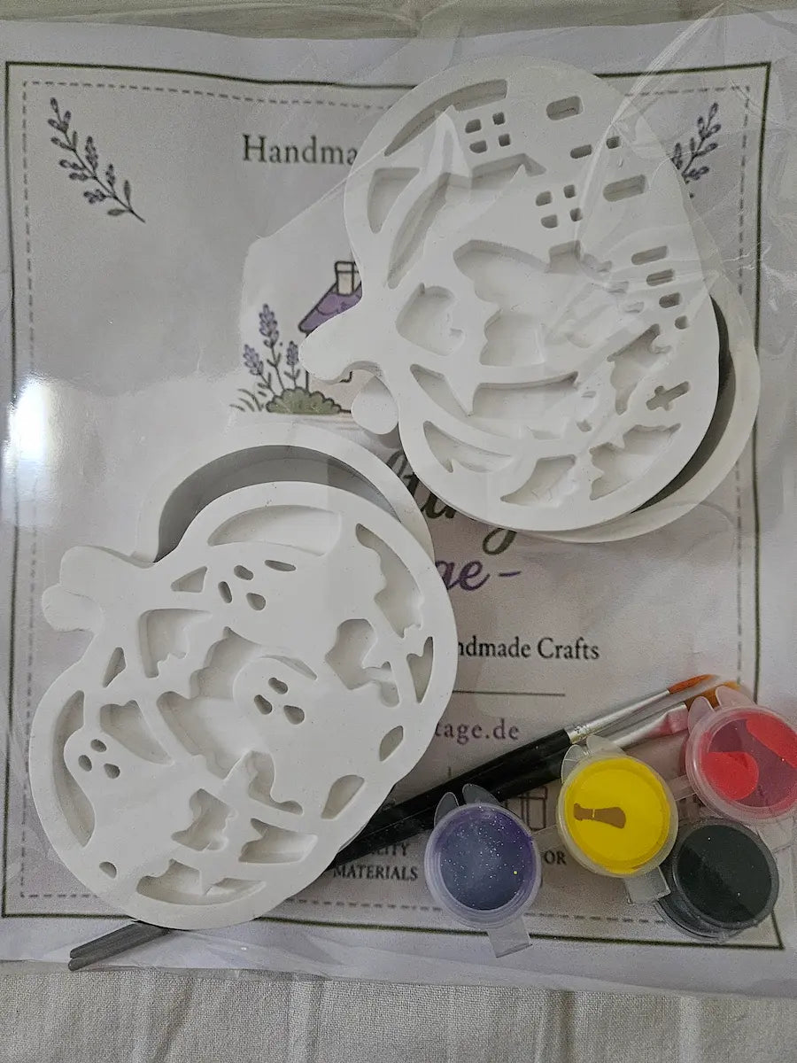 A Halloween-themed craft kit featuring two white plastic pumpkin-shaped cookie cutters, a paintbrush, and three small paint pots in purple, yellow, and red, all laid out on a printed white craft instruction sheet.