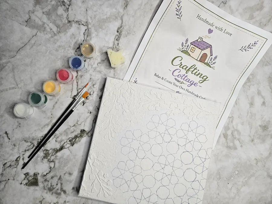 A flat-lay of a DIY craft kit on a gray marble surface, featuring a printed instruction sheet for 'Crafting Cottage', a white canvas with geometric line patterns, two paint pots, and two paintbrushes.