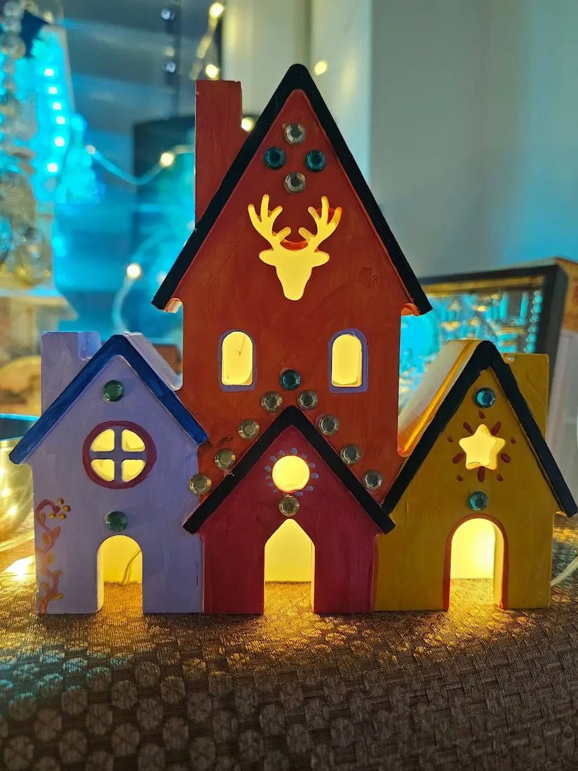 A set of three hand-painted wooden Christmas houses with warm white LED lights glowing from within, decorated with colorful gems and festive cutouts like a reindeer and a star.