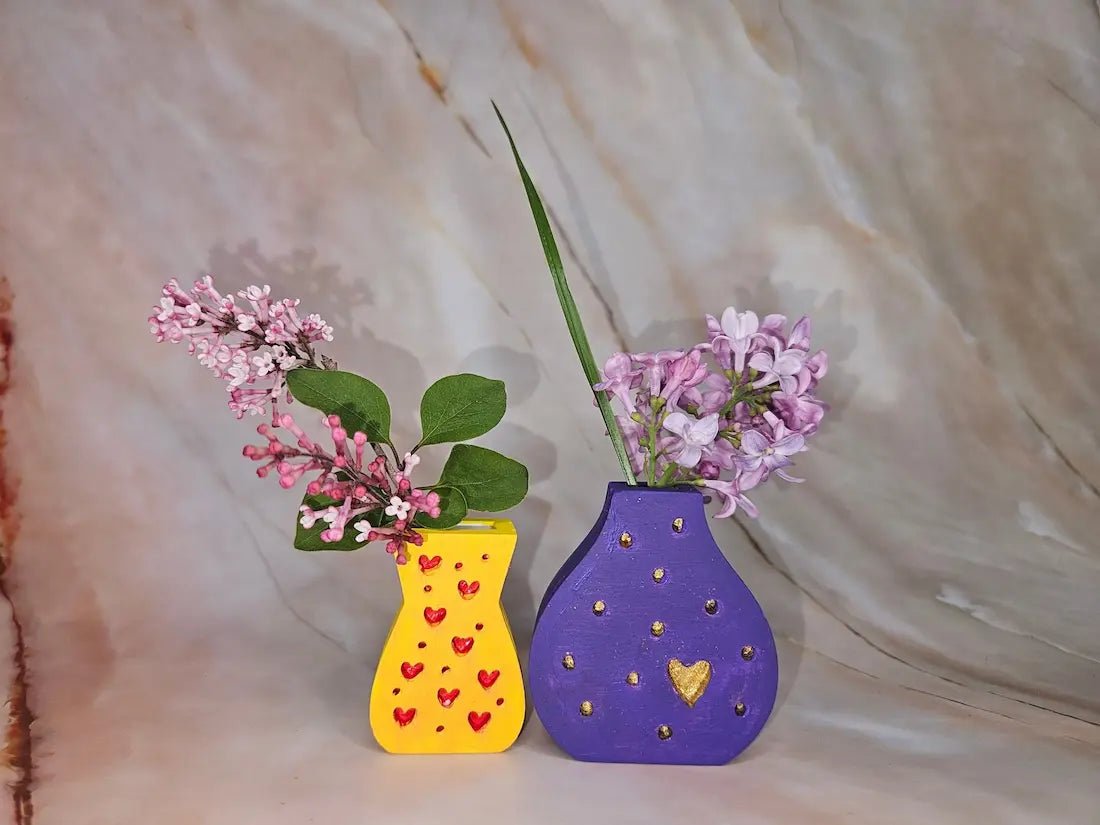 Two decorative wooden vases, one yellow with red hearts and one purple with gold dots and a gold heart, holding pink and purple flowers, set against a beige marble-patterned backdrop.